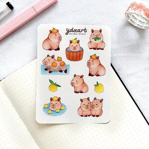 Peut inclure: Une feuille d'autocollants représentant d'adorables capybaras en dessin animé savourant des oranges. Les autocollants sont sur un fond blanc avec le texte "ydxcart" et "Sticker Sheets".