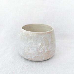 May include: A small, round ceramic cup with a textured, off-white exterior. The cup has a wide opening and a slightly pinkish hue near the base. The cup is likely handmade and suitable for beverages.