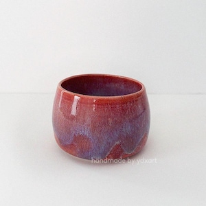 Peut inclure: Un bol en céramique fait à la main de forme arrondie. Le bol présente une finition brillante avec un mélange de teintes rouges et violettes. Les mots "handmade by yoxart" sont imprimés sur le côté du bol.