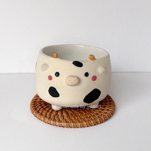 May include: A cream-coloured ceramic cow-shaped planter with black spots, pink cheeks, and small horns. The planter sits on a round, woven coaster. A decorative item for home decor.