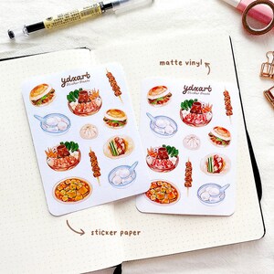 May include: Two sheets of stickers featuring illustrations of various Chinese food dishes. The top sheet is labeled "matte vinyl" and the bottom sheet is labeled "sticker paper". The stickers include a bowl of soup, a plate of dumplings, a plate of noodles, a plate of meat skewers, and a plate of fried tofu.