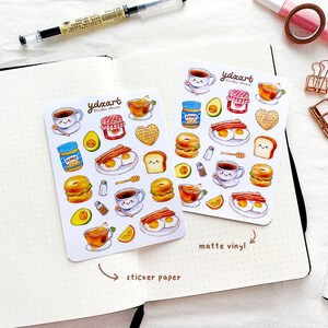 May include: Two sheets of stickers featuring a breakfast theme. The stickers are on white paper with a white background. The stickers are printed with a variety of breakfast foods, including eggs, bacon, toast, coffee, and avocado. The stickers are labeled "ydncart sticker sheets" and "matte vinyl".