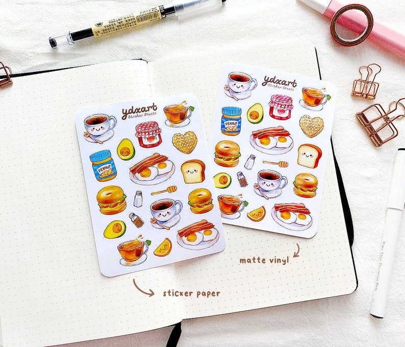 May include: Two sheets of stickers featuring a breakfast theme. The stickers are on white paper with a white background. The stickers are printed with a variety of breakfast foods, including eggs, bacon, toast, coffee, and avocado. The stickers are labeled "ydncart sticker sheets" and "matte vinyl".