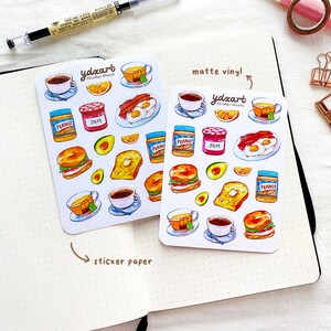 May include: Two sheets of stickers featuring breakfast food illustrations. The top sheet is labeled "matte vinyl!" and the bottom sheet is labeled "sticker paper". Both sheets feature illustrations of coffee, tea, toast, eggs, bacon, peanut butter, jam, avocado, and bagels.