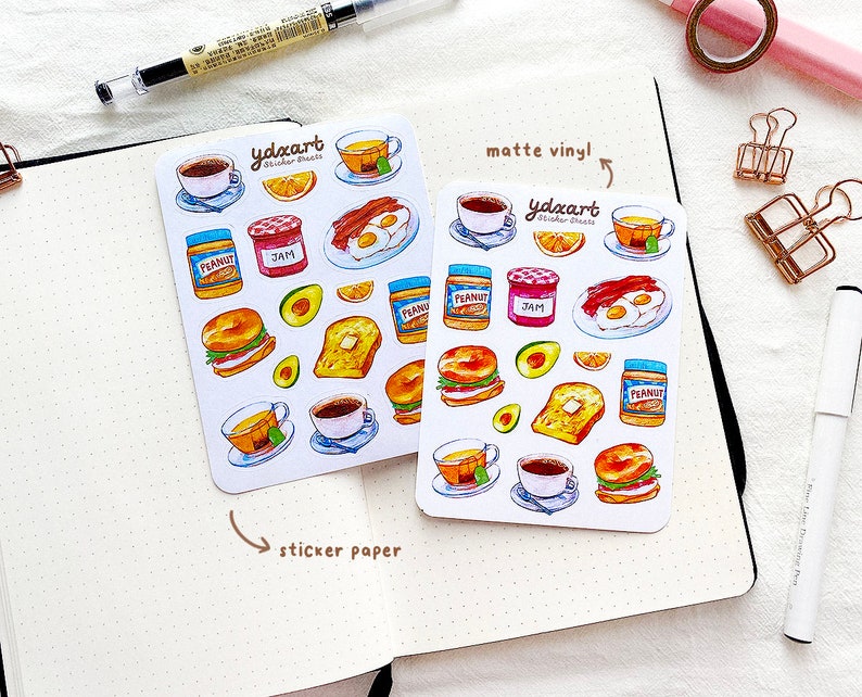May include: Two sheets of stickers featuring breakfast food illustrations. The top sheet is labeled "matte vinyl!" and the bottom sheet is labeled "sticker paper". Both sheets feature illustrations of coffee, tea, toast, eggs, bacon, peanut butter, jam, avocado, and bagels.
