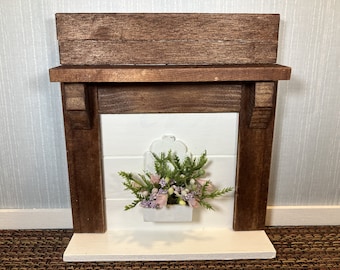 May include: A miniature wooden fireplace mantel painted white and stained brown. The mantel features a decorative floral arrangement in a white planter box. The overall design is rustic and charming, suitable for home decor.
