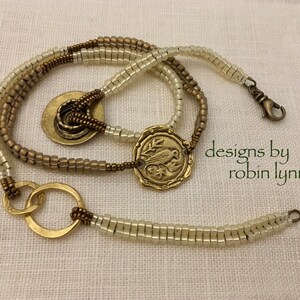 May include: A multi-strand necklace with gold and cream beads, featuring a large circular pendant with a bird design and the text "designs by robin lynn".