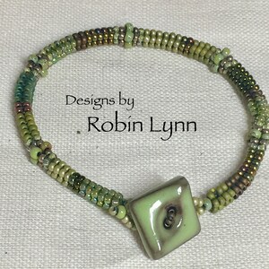 May include: A green and brown beaded bracelet with a square ceramic button in the center. The button has a light green glaze and a brown swirl design. The bracelet is made with seed beads and has a delicate, feminine look. Designs by Robin Lynn.