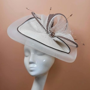 May include: Elegant white and brown formal hat. The hat has a wide brim with a black trim, a patterned ribbon, and delicate netting. Small, thin, brown embellishments are also present. A stylish accessory for special occasions.