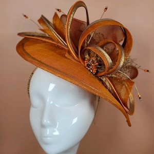 May include: A striking orange and gold fascinator. The headpiece features a wide brim and a layered floral design. It includes spiraling ribbons, gold accents, and beaded embellishments. The hat is displayed on a white mannequin head, showcasing its elegant design.