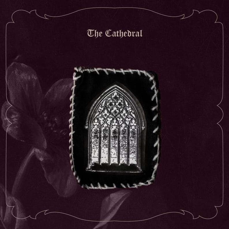 May include: A black and white pin featuring a cathedral window design, set against a black background with white stitching. The pin is displayed on a dark purple background with the text "The Cathedral".