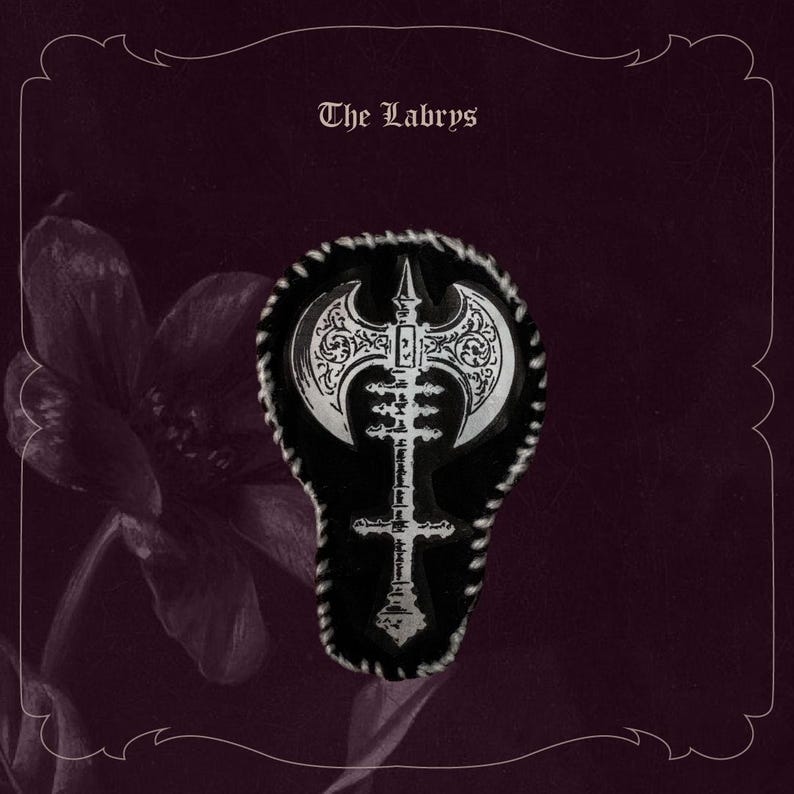 May include: A silver and black pin with an axe design, set against a black background with a decorative border. The pin is shaped like an axe and has intricate patterns. The text "The Labrys" is at the top.