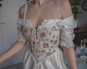 Könnte beinhalten: Ein weißes, schulterfreies Kleid mit einem floralen Korsett-Oberteil und Spitzenbesatz. Das Kleid hat ein figurbetontes Oberteil und einen fließenden Rock.
