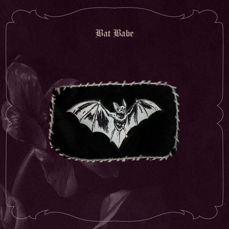 May include: A black rectangular patch featuring a white bat design and the text "Bat Babe". The patch is stitched with white thread and set against a dark burgundy background with a floral element.