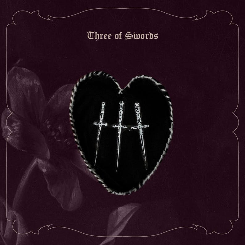 May include: A tarot card illustration with a black heart shape pierced by three silver swords. The card is titled "Three of Swords" in a decorative font, set against a dark burgundy background with floral accents and a decorative border.