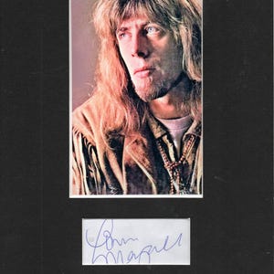 May include: A black and white photo of a man with long, blonde hair. He is wearing a brown leather jacket and a beaded necklace. The photo is signed "John Mayall".