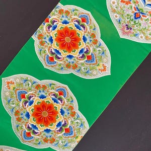 May include: Green fabric with three embroidered floral designs. Each design features a central flower with multiple layers of petals in shades of red, orange, yellow, blue, and white. The flowers are surrounded by smaller flowers and leaves in shades of green, white, and pink.