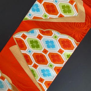 Op de afbeelding: Een rode stof met een geometrisch patroon in wit, oranje, groen en blauw. Het patroon heeft een herhalend ontwerp van achthoeken en vierkanten met bloemmotieven.