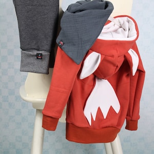 May include: A children's outfit comprising a red fox-themed hoodie with white details and a pair of grey leggings. The hoodie has a hood with fox ears and a white tail design on the back. A grey scarf is also included.