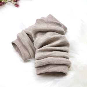 Könnte beinhalten: Ein Paar beige Beinwärmer aus weichem Rippstrick. Die Beinwärmer sind zerknittert, wodurch die Textur des Materials sichtbar wird. Sie werden auf einer weißen, flauschigen Oberfläche mit einem Zweig Beeren im Hintergrund präsentiert.