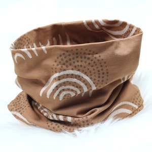 Op de afbeelding: Een bruine stoffen nekwarmer met een wit en bruin geometrisch patroon. Het patroon bevat cirkels en bogen.