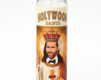 May include: A white candle with a gold label that reads "Hollywood Saints" and a picture of Ryan Reynolds dressed as Jesus with a crown and a heart on his chest. The label at the bottom reads "St Ryan".