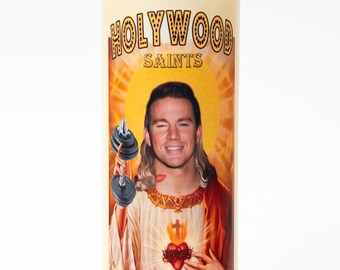 May include: A glass candle with a yellow, orange, and red image of a man with long blonde hair holding a dumbbell. The image is surrounded by roses and doves. The text "HOLLYWOOD SAINTS" is at the top of the image and "ST CHANNINNG" is at the bottom.