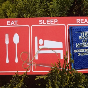 May include: A red sign with three sections, each with a white icon and text. The first section says "EAT." and has a fork and spoon. The second section says "SLEEP." and has a bed with a person sleeping in it. The third section says "READ." and has a blue book with the text "THE BOOK OF MORMON ANOTHER TESTAMENT OF JESUS CHRIST" on it.