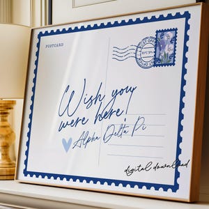 May include: A framed print resembling a postcard, featuring the text "Wish you were here!" in elegant script. The design includes a blue postage stamp and border, with the words "Alpha Delta Pi" and "digital download" also visible.