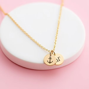 May include: A gold-coloured necklace featuring a delicate chain and two small, circular pendants. One pendant is engraved with a black anchor symbol, while the other pendant displays the Greek letters Delta and Gamma in black.