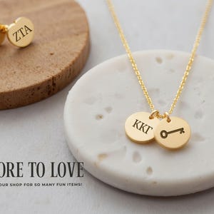 May include: Gold-colored jewelry set featuring a necklace with two round pendants and a pair of stud earrings. The necklace pendants display initials and a key symbol. The earrings also have initials. The jewelry is displayed on a white and brown surface.