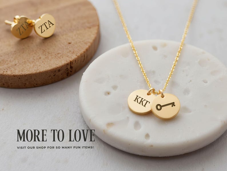 May include: Gold-colored jewelry set featuring a necklace with two round pendants and a pair of stud earrings. The necklace pendants display initials and a key symbol. The earrings also have initials. The jewelry is displayed on a white and brown surface.