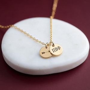 May include: A gold-toned necklace featuring a delicate chain and two small circular pendants. One pendant is engraved with a horizontal arrow, and the other pendant displays the Greek letters Pi Beta Phi, represented as ΠΒΦ.