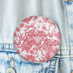 May include: A round button with a white background and a red floral and bow pattern. The words "Gamma Phi Beta" are written in a cursive font. The button is pinned to a denim jacket.