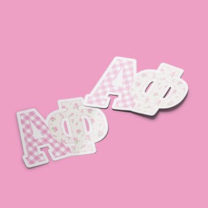 May include: Two decorative letter cutouts, "A" and "Φ", in a pink and white gingham pattern and a floral pattern. The letters are white with a pink background. The background is a solid pink color.