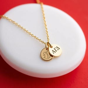 May include: A gold-coloured necklace featuring a delicate chain and two small circular pendants. One pendant displays an engraved squirrel silhouette, while the other pendant features the black Greek letters Alpha Gamma Delta, or ΑΓΔ.