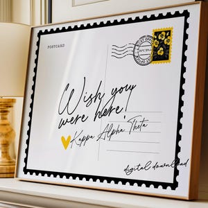 May include: A framed postcard print with a black border and a wooden frame. The postcard reads "Wish you were here!" in cursive, with a yellow heart and the text "Kappa Alpha Theta." A postage stamp with yellow flowers is in the upper right corner.