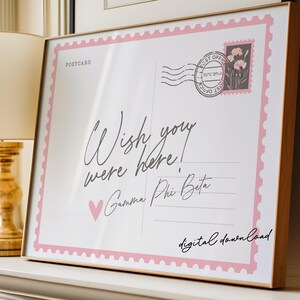 May include: A framed print resembling a postcard, featuring the text "Wish you were here!" in elegant script. The design includes a pink border, a heart, and a postage stamp graphic. The words "Gamma Phi Beta" and "digital download" are also present.