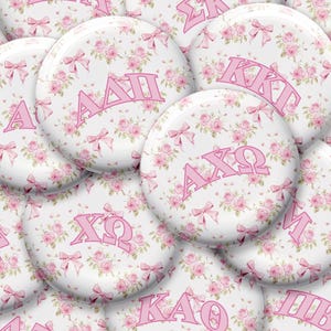 May include: A collection of round, white buttons with pink floral patterns and pink bows. Each button features Greek letters in pink, representing sororities. The buttons are scattered, creating a visually appealing arrangement.