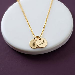 May include: A gold-toned chain necklace featuring two small circular pendants. One pendant displays an engraved sailboat silhouette, while the other pendant features the Greek letters Sigma Sigma Sigma engraved in a clear, dark font.