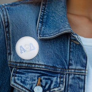 May include: A circular pinback button featuring a white background with subtle vertical stripes. The centre displays the Greek letters Alpha Xi Delta in a light blue and white checkered pattern with a distressed, textured appearance.