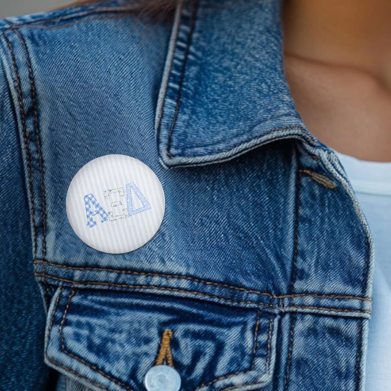 May include: A circular pinback button featuring a white background with subtle vertical stripes. The centre displays the Greek letters Alpha Xi Delta in a light blue and white checkered pattern with a distressed, textured appearance.