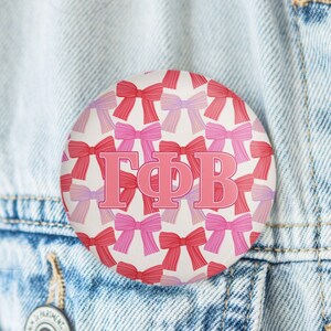 May include: A round button with a white background, covered in a pattern of pink and red bows. The Greek letters ΓΦΒ are in the center of the button, in a pink color. The button is pinned to a denim jacket.