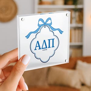 May include: A clear acrylic block featuring a white design with blue accents. The design includes a blue bow, a decorative frame, and the Greek letters &Alpha;&Delta;&Pi;. The text "BE THE FIRST" is also included.