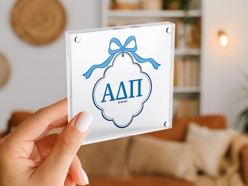 May include: A clear acrylic block featuring a white design with blue accents. The design includes a blue bow, a decorative frame, and the Greek letters &Alpha;&Delta;&Pi;. The text "BE THE FIRST" is also included.