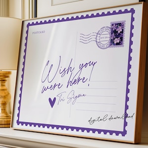 May include: A framed print designed to resemble a postcard. The white background features purple text that reads "Wish you were here!" and "Tri Sigma." A purple stamp and border complete the design.
