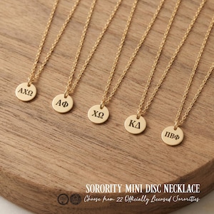 May include: Five gold-toned necklaces with small circular pendants, each engraved with different Greek letters: ΑΧΩ, ΑΦ, ΧΩ, ΚΔ, and ΠΒΦ. The delicate chains are attached to the round discs, which feature a polished finish and dark lettering.