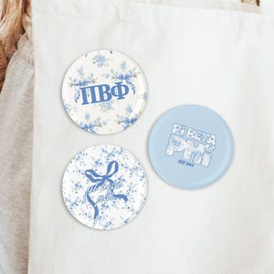 May include: Three round buttons with blue floral designs and text. One button has Greek letters, another has a bow and script, and the third has "Pi Beta Phi" and "EST 1867". The buttons are on a white background.