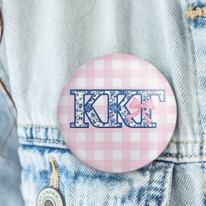 May include: A round button with a pink and white gingham pattern background. The button features the letters "KKT" in a blue floral design, with a pink bow. The button is pinned to a denim jacket.