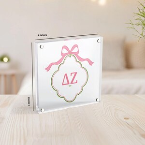 Puede incluir: Un bloque acr&iacute;lico transparente, de aproximadamente 10 cm cuadrados, con un lazo rosa y un emblema decorativo con las letras griegas &Delta;Z en rosa. El emblema est&aacute; delineado en verde y rosa. El bloque se muestra sobre una superficie de madera clara.
